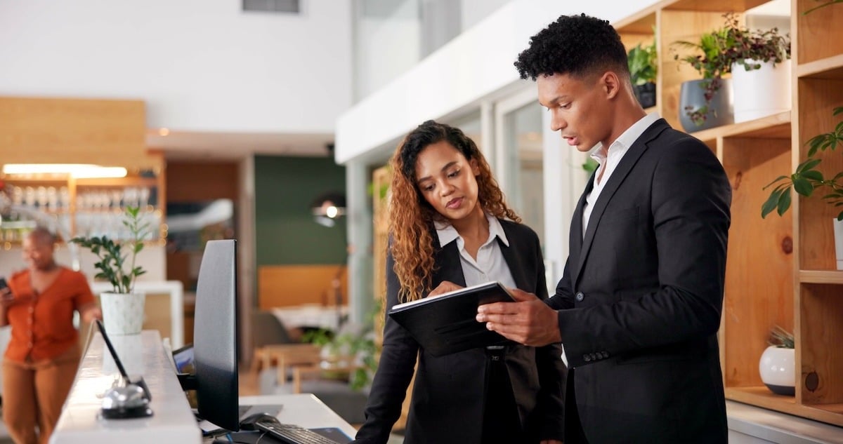 Two hospitality professionals standing behind a front desk and looking at information on a tablet. 