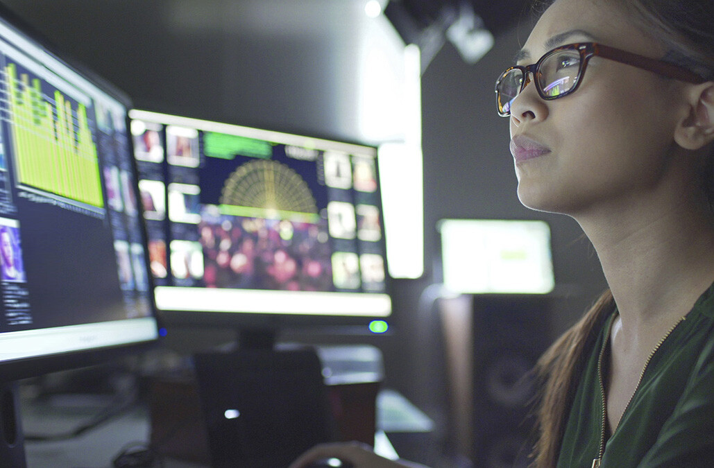 Close up stock image of a young asian woman sitting down at her desk where she’s surrounded by 3 large computer monitors displaying out of focus images of people as