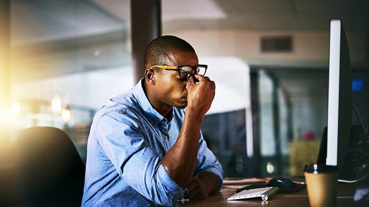 an image of a male employee pinching his nose bridge while sitting in front of a computer with eyes closed