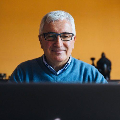 an older man with grey hair and blue sweater watching his laptop smiling