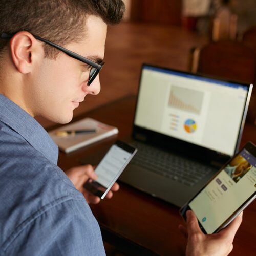 Attractive man in glasses working with multiple electronic internet devices. Freelancer businessman has laptop and smartphone in hands and laptop on table with charts on screen. Multitasking theme