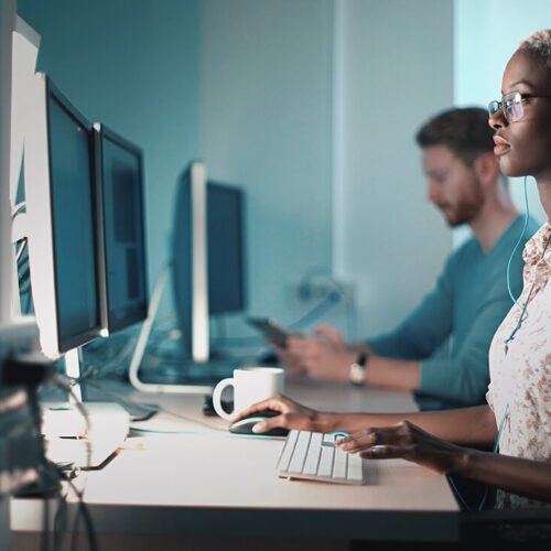 a woman with one hand on the mouse and another typing on the keyboard while watching the monitor in front of her with a male colleague to her right out of focus